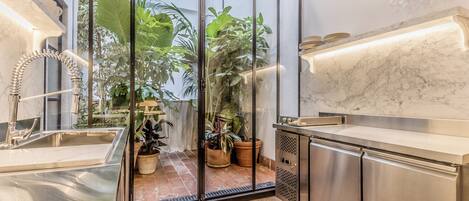 A modern kitchen with a stainless steel sink, a marble countertop, and a built-in oven. There are potted plants and a view of greenery outside.