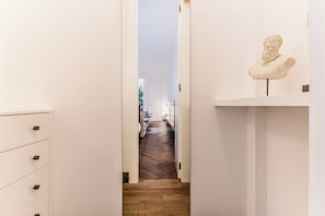 Un pasillo con piso de madera, pared blanca y un busto sobre un pedestal.