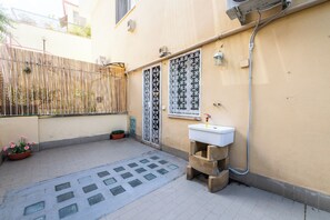 Um pequeno pátio com piso de azulejos, cerca de metal, planta em vaso, pia e janela com detalhes em metal decorativo.
