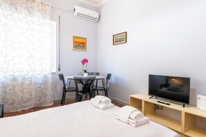 Ein Hotelzimmer mit Bett, Fernseher, Schreibtisch mit Stuhl, Tisch mit Pflanze und zwei gerahmten Bildern an der Wand.