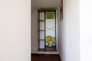 Un estrecho pasillo con un estante metálico alto y negro contra la pared, que incluye un jarrón con flores y un cuadro enmarcado encima.