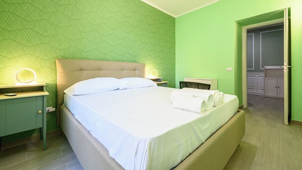 Un lit bien fait dans une pièce aux murs verts et ornée d’un motif géométrique. On y trouve une table de chevet avec une lampe et un petit miroir. Une porte donne accès à une salle de bain avec un lavabo et un meuble blancs.
