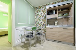 Een compacte keuken met een magnetron, spoelbak en houten kastjes. Er is een klein eethoekje met een glazen tafel en stoelen.