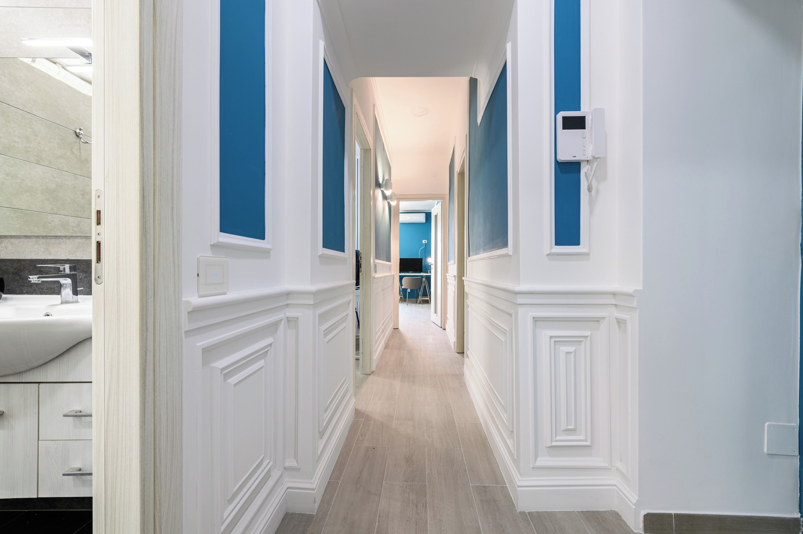 A hallway with white wainscoting and blue walls, leading to a room with a large window.