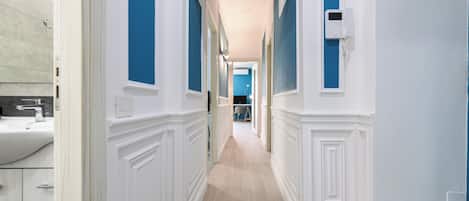 A hallway with white wainscoting and blue walls, leading to a room with a large window.
