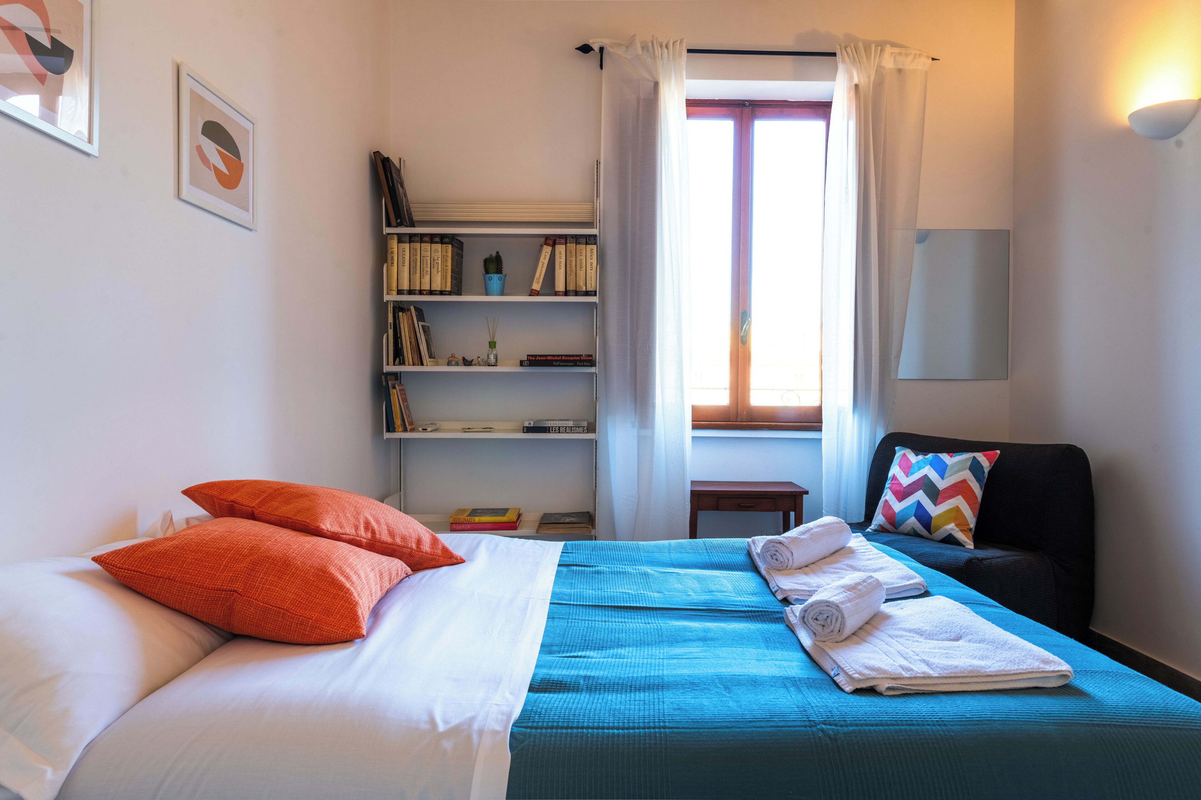 A neatly arranged bedroom with a bed, pillows, a blue bedspread, a bedside table with a lamp, a bookshelf, and a window with curtains.