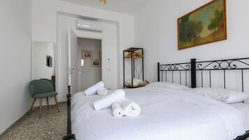 Un letto rifatto con lenzuola bianche e asciugamani arrotolati, una poltrona verde, un quadro appeso al muro e un lampadario al soffitto.