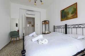 A neatly made bed with white linens and rolled towels, a green chair, a painting on the wall, and a ceiling light fixture. - Via Coriolano 8 (Naples)