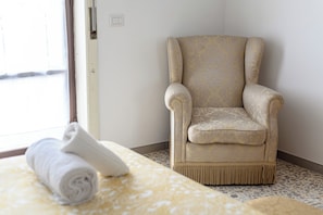 Quarto com colcha estampada, toalha branca enrolada e poltrona bege com tecido estampado.