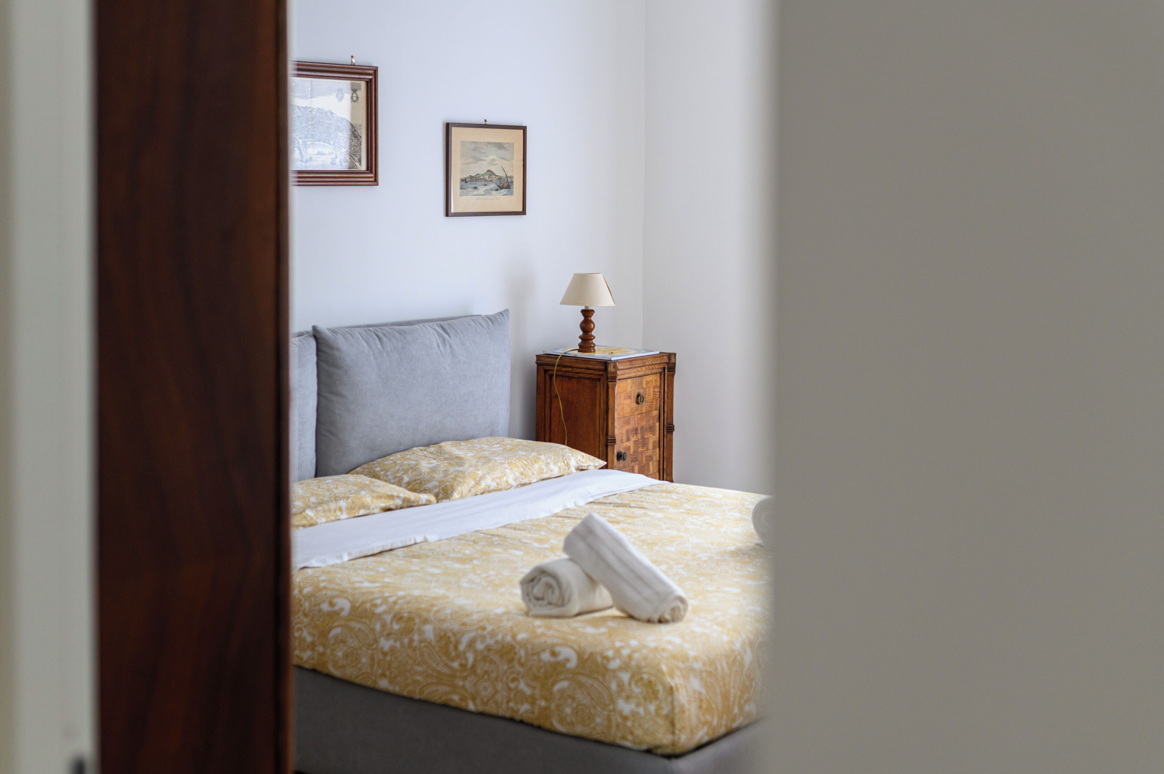 Een opgemaakt bed met een bedsprei en kussens, een nachtkastje met een lamp, en twee ingelijste foto's aan de muur.