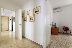 Un couloir avec une porte donnant sur une pièce, une petite armoire en bois sur laquelle se trouve un téléviseur, et une chaise rouge.