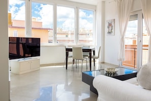Ein modernes Wohnzimmer mit Sofa, Glastisch, Fernseher und einem Essbereich mit Stühlen und Tisch.
