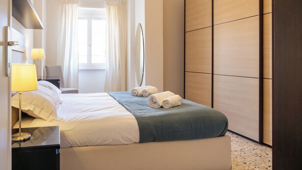 Una camera d'albergo con un letto, un comodino con una lampada, un armadio e una finestra con tende.