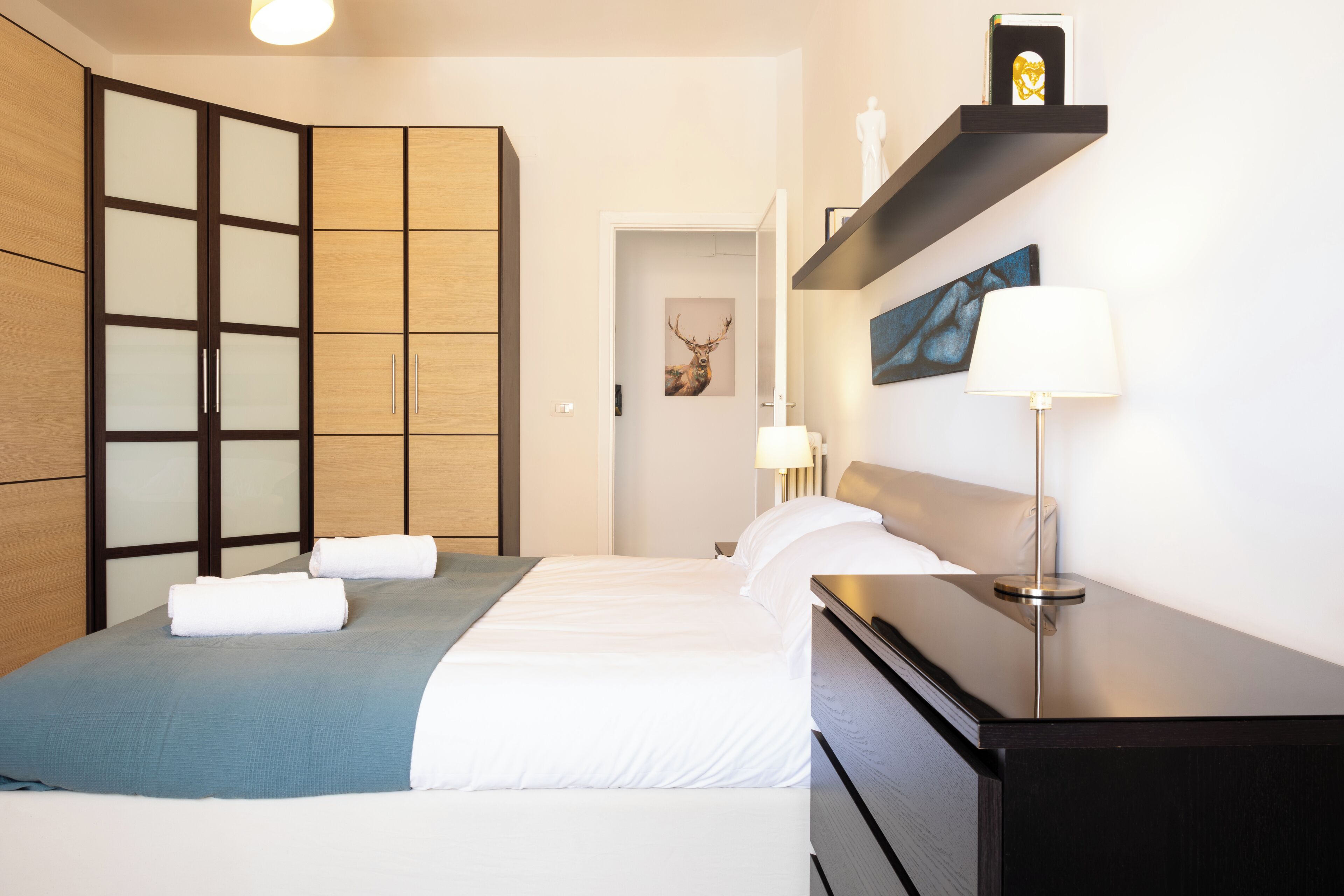 A bedroom with a bed, a dresser, a nightstand, a lamp, a shelf with a picture, and a wardrobe.