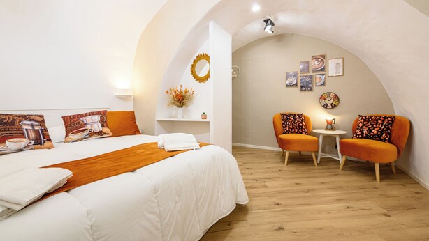 Quarto de hotel com cama, uma mesinha, duas cadeiras, parede com quadros e um objeto decorativo na parede.