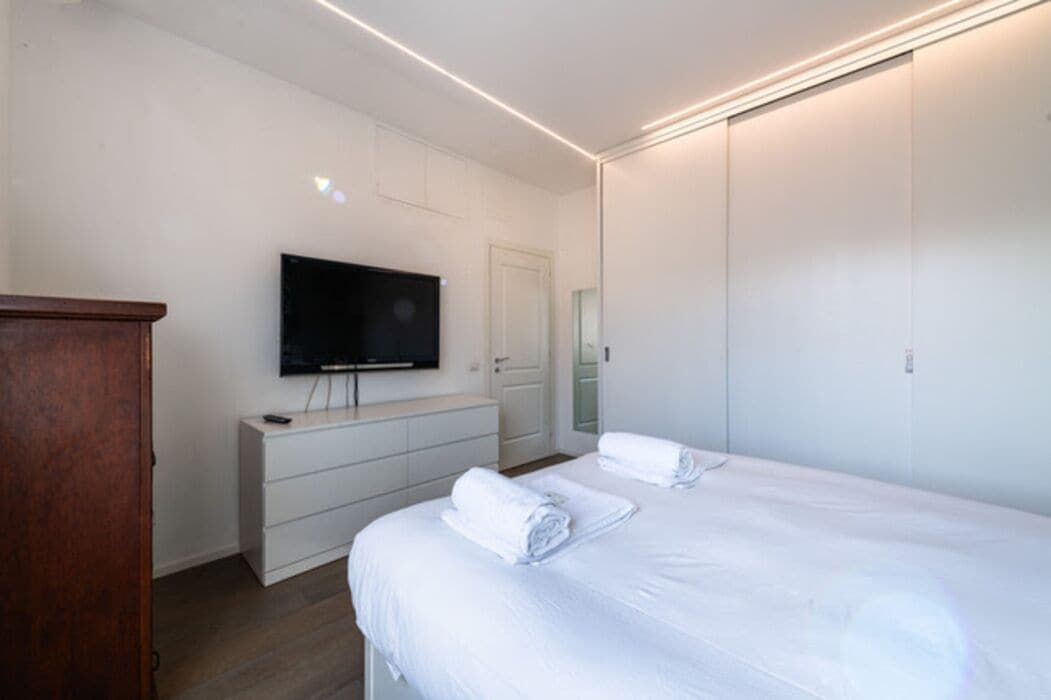 Quarto moderno com cama grande, televisão na parede, cómoda e guarda-roupa.