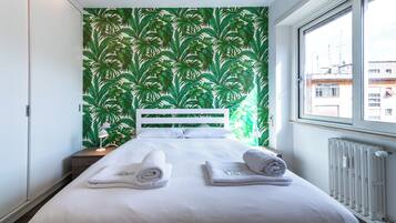 A hotel room with a bed, white headboard, and a green leafy wallpaper.