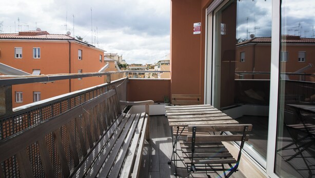 En balkon med et træbord og stole, et metalgelænder og udsigt til omkringliggende bygninger.