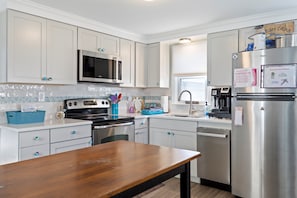 A modern kitchen with white cabinets, stainless steel appliances, and a wooden table. - Beach bungalow |Fenced yard |Steps to toes in sand (Surf City)