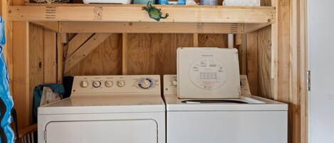 A laundry area with two white washing machines, a wooden shelf above with various items, and a blue towel on the left.