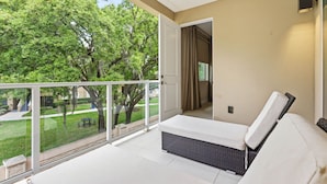 A balcony with a white sofa, a wicker chair, and a view of a green lawn and trees.