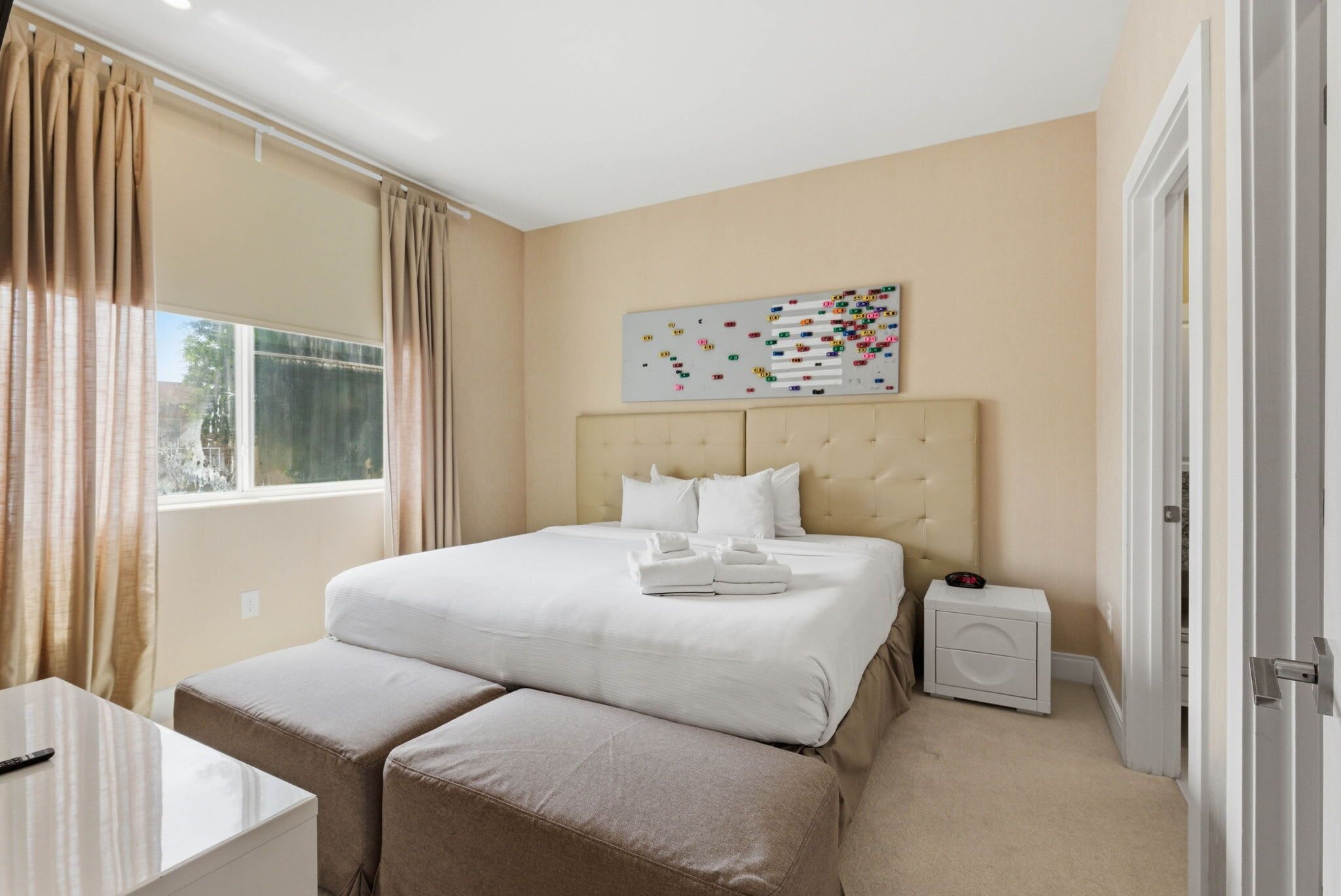 Une chambre d’hôtel comprenant un lit, une table de chevet, un banc, une commode, une œuvre d’art murale et une fenêtre avec des rideaux.