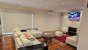 Een moderne woonkamer met een hoekbank, een glazen salontafel, een witte poef, een open haard en een televisie.
