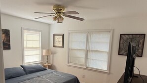 Une chambre à coucher avec un lit, un ventilateur de plafond, une fenêtre avec des stores, une lampe et un téléviseur posé sur un meuble.