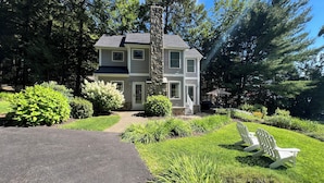 A two-story house with a stone chimney, surrounded by lush greenery and a paved driveway.