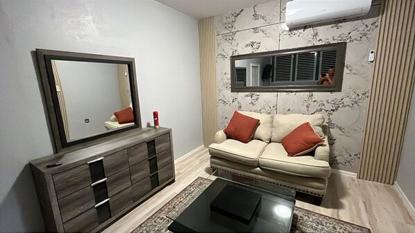 Una sala de estar con sofá, mesa de centro, cómoda, espejo y aire acondicionado instalado en la pared.