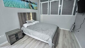 A bedroom with a bed, a nightstand, a wall-mounted TV, and a large window with shutters. - Beautiful Hideaway with pool close to hiking, waterfalls and Culebra ferry (Ceiba)