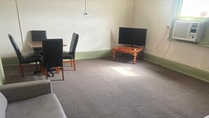 A room with a grey carpet, a TV on a wooden stand, a table with chairs, and a window with an air conditioner.
