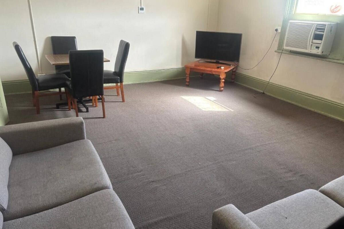 Une pièce avec un tapis gris, un téléviseur posé sur un meuble en bois, une table avec des chaises et une fenêtre avec un climatiseur.