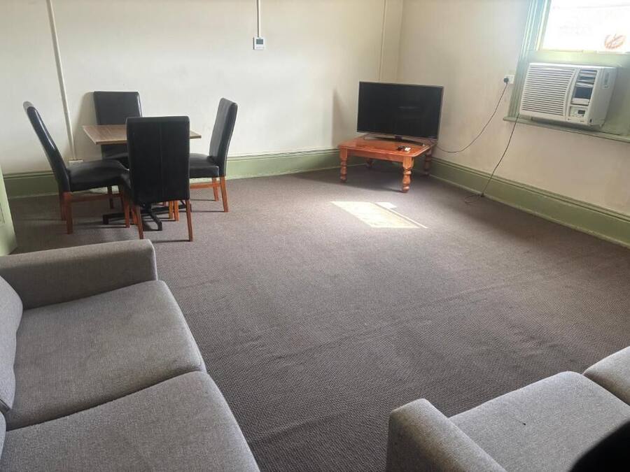 A room with a grey carpet, a TV on a wooden stand, a table with chairs, and a window with an air conditioner.