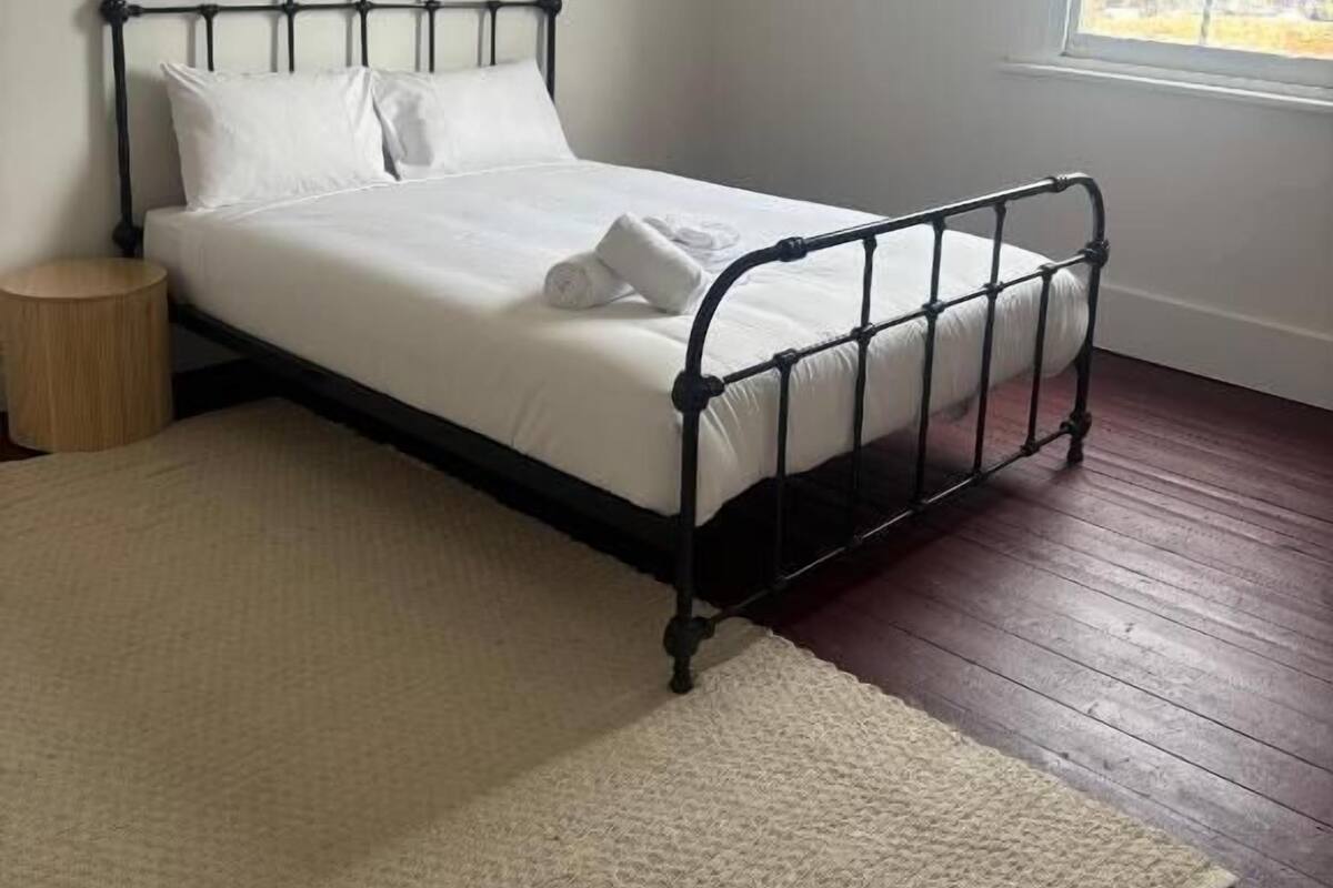 A single bed with a metal frame, a bedside table, a window with a view, and a carpeted floor.