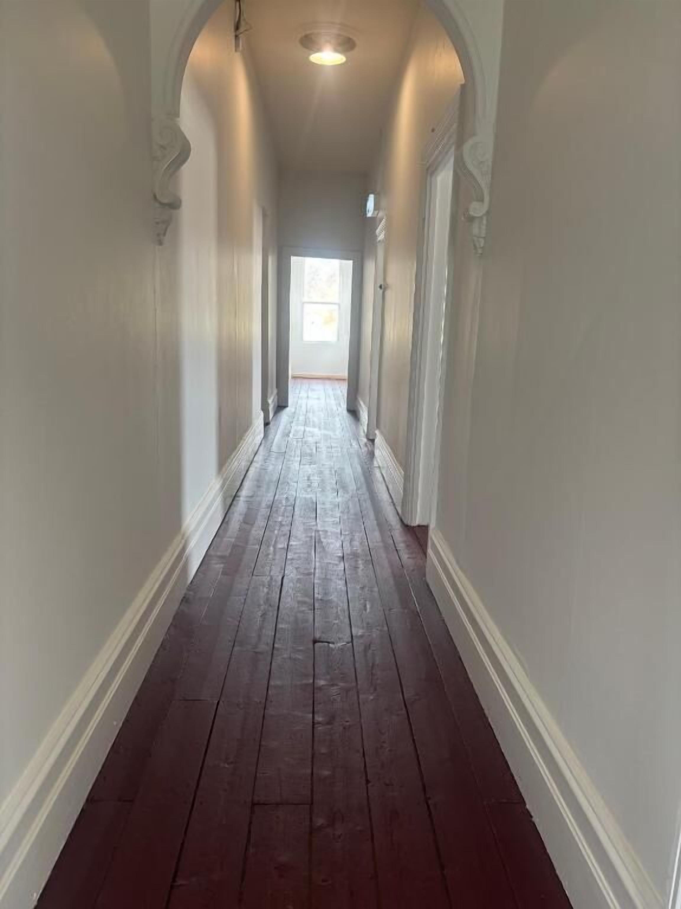 A long, narrow hallway with a wooden floor and white walls, leading to a door at the end.