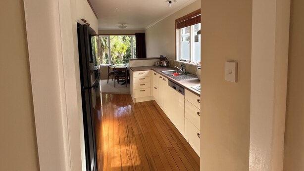 Una cocina con piso de madera, armarios blancos, un ventanal con vista a follaje y una zona de comedor con mesa y sillas.