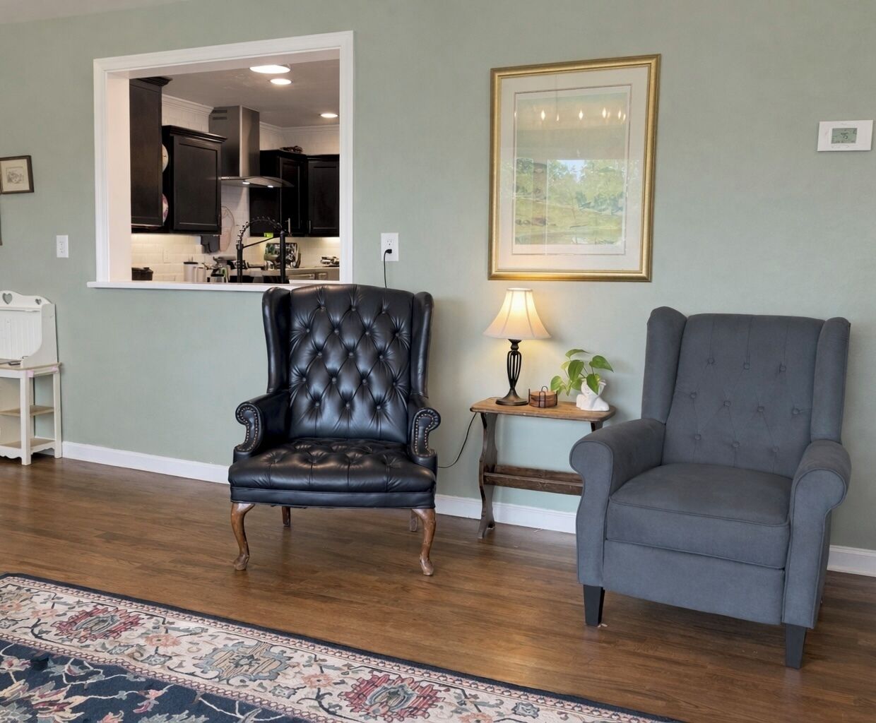 A living room with a dark leather armchair, a grey armchair, a wooden side table with a lamp, and a framed picture on the wall.