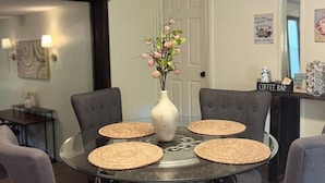 A dining area with a round glass table, four grey upholstered chairs, a white vase with pink flowers, and a coffee bar sign. - Arbuckle Lake Hideaway (Sulphur)