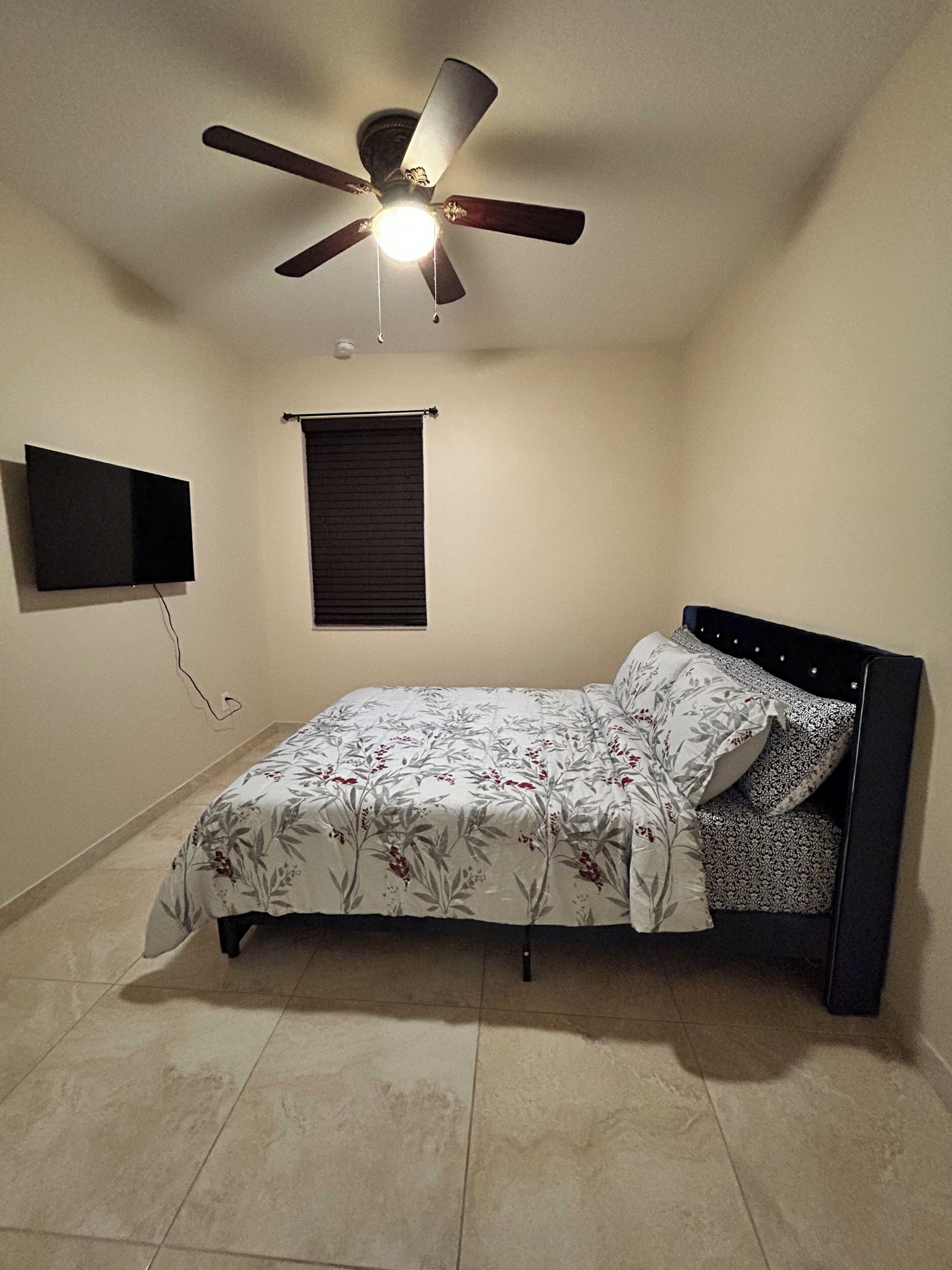Un dormitorio con cama, ventilador de techo, televisor montado en la pared y ventana con persianas.