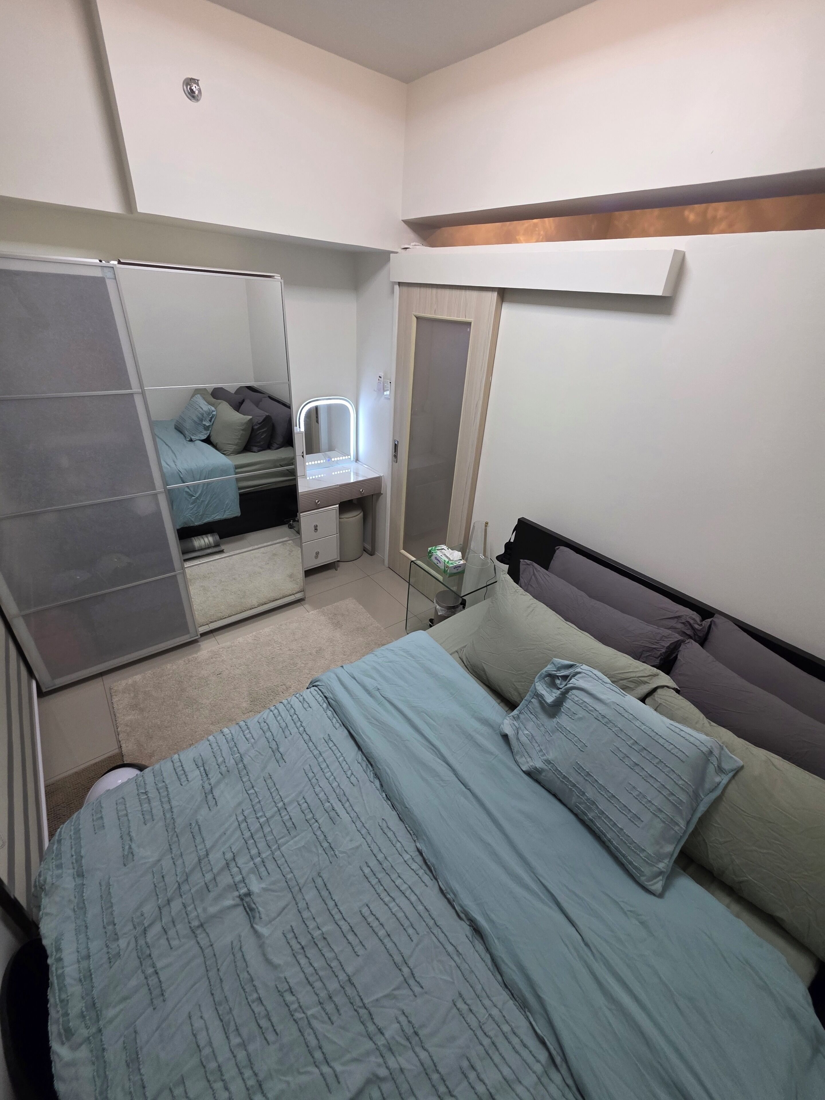 A bedroom with a bed, a vanity with a mirror, a wardrobe, and a small table with a lamp.