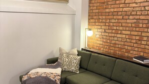 Een woonkamer met een groene bank, twee voetenbankjes, een gebogen plaid, een lamp en een bakstenen muur.