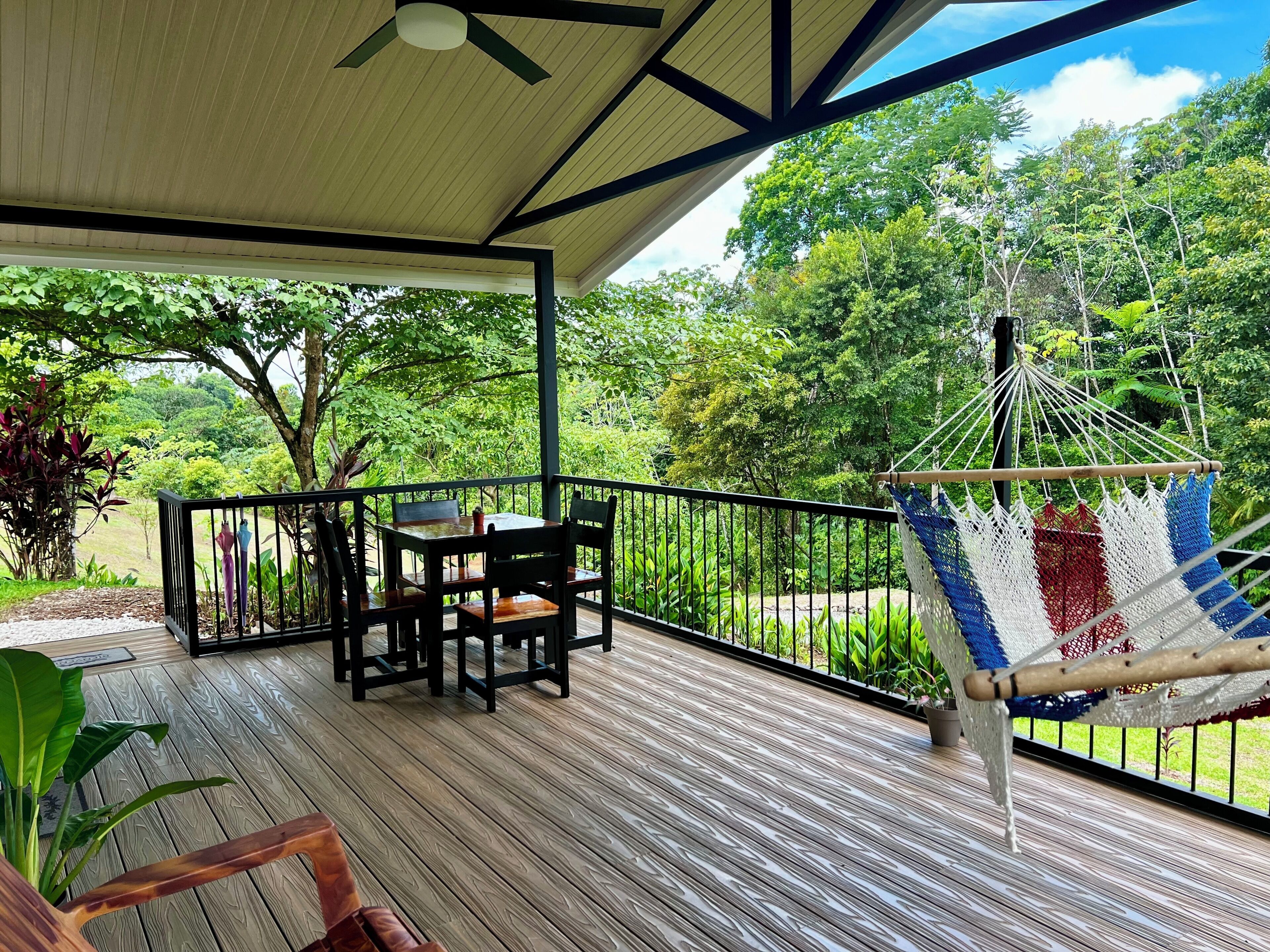 A wooden deck with a table and chairs, a hammock, and a view of lush greenery.