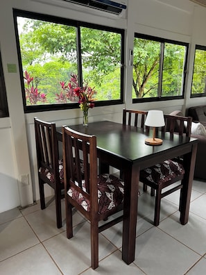 A dining area with a table, chairs, a vase with flowers, and large windows with a view of greenery.