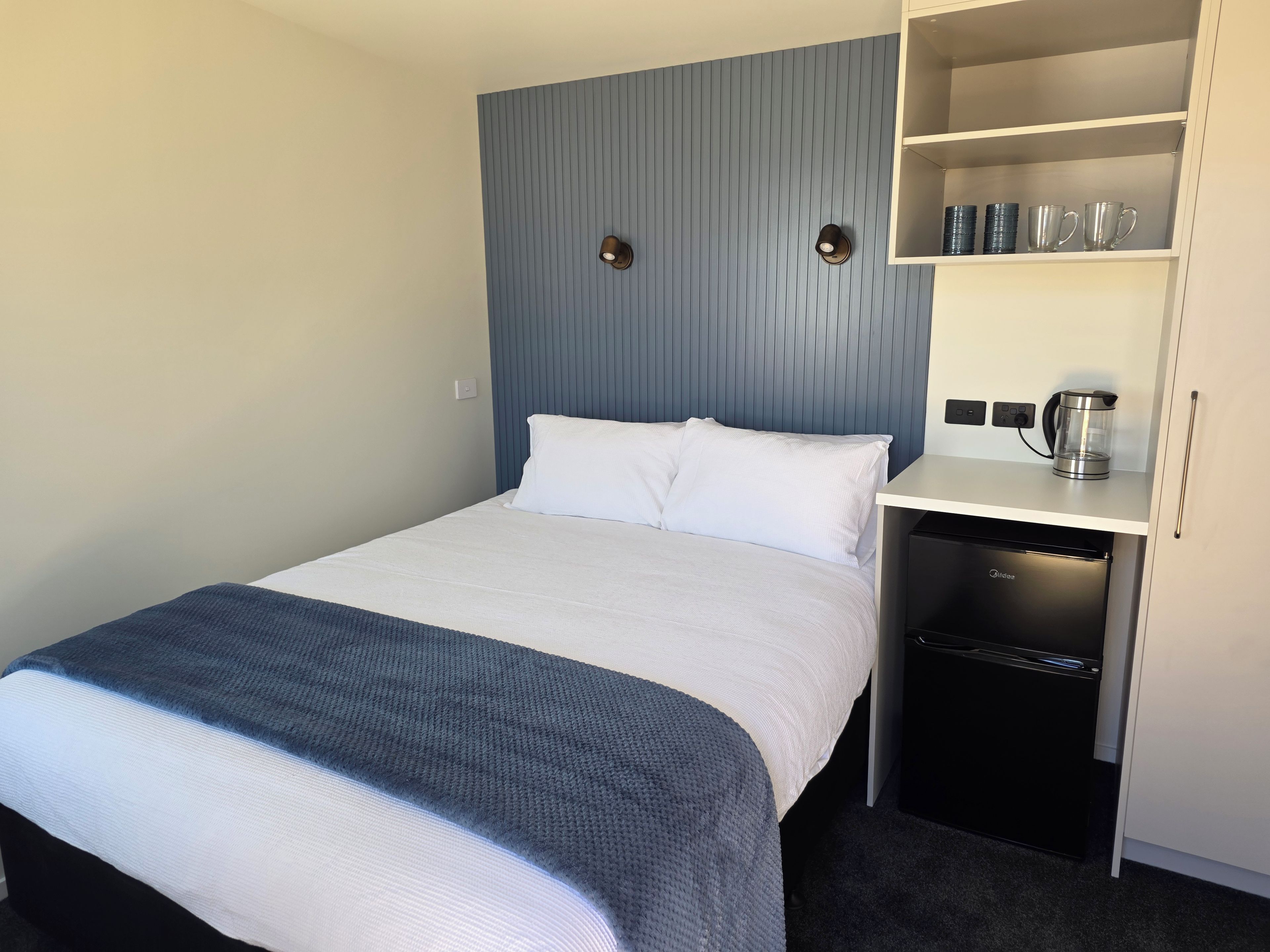 A hotel room with a bed, a coffee maker, a shelf with cups, and a black appliance.