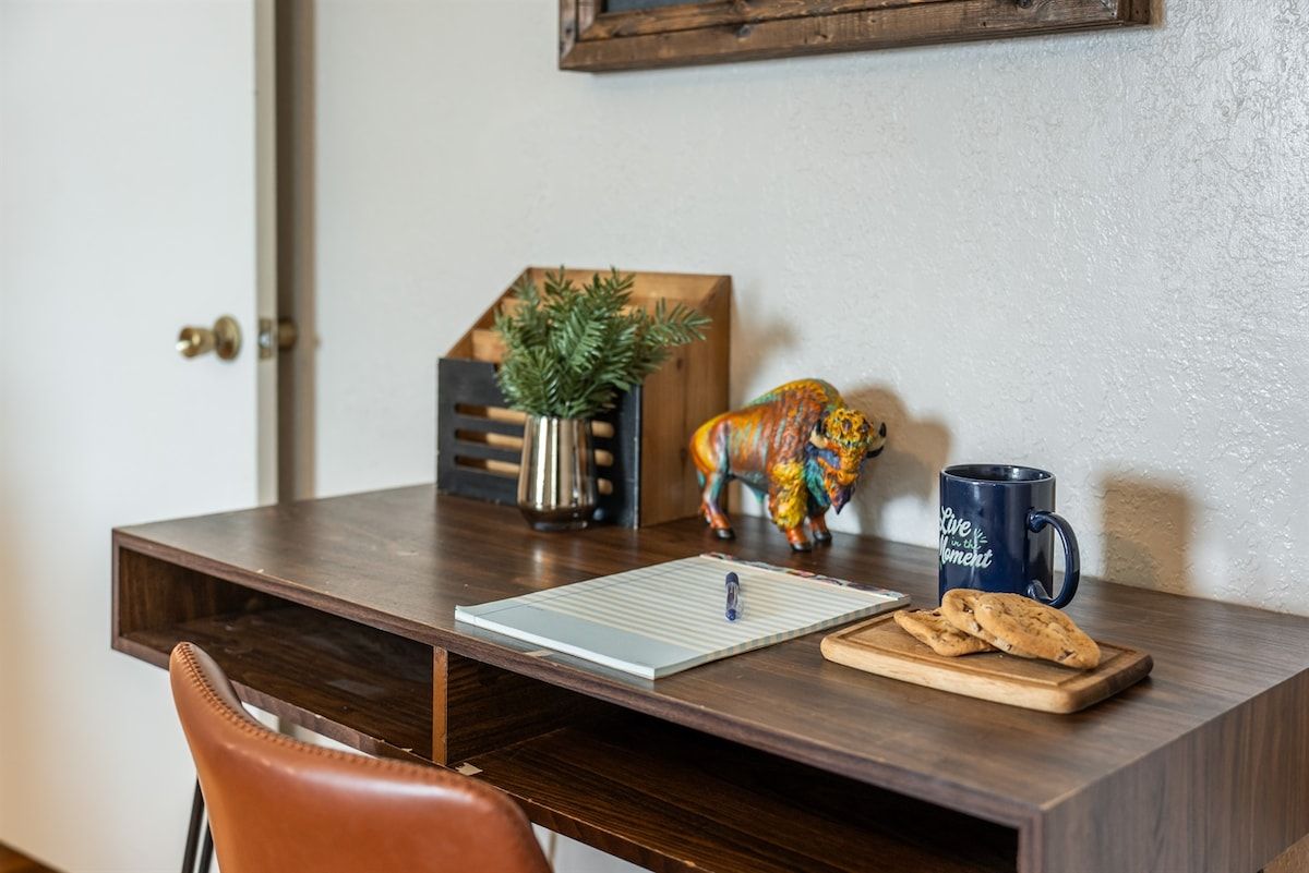 A wooden desk with a notepad, pen, mug, cookies, a plant, and a colorful figurine.