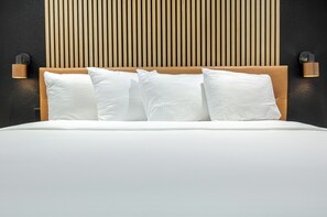 Cama com cabeceira de madeira, roupa de cama branca e duas luminárias fixadas na parede.