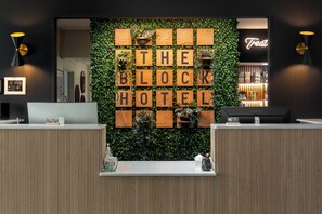 Uma mesa de recepção com uma parede de blocos de madeira exibindo "THE BLOCK HOTEL" e um letreiro de neon com a palavra "Treat".