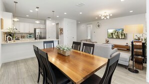 A modern kitchen with white cabinets, a dining table, and a living area with a sofa and TV. - Game Day Stay - Community Pool Access + Walk to Stadium + Close to Westgate (Glendale)