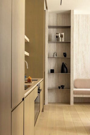 Eine moderne Küche mit Einbauofen, Spüle und einer Arbeitsplatte mit Toaster. Regale mit Dekoration und eine Bank im angrenzenden Wohnbereich.